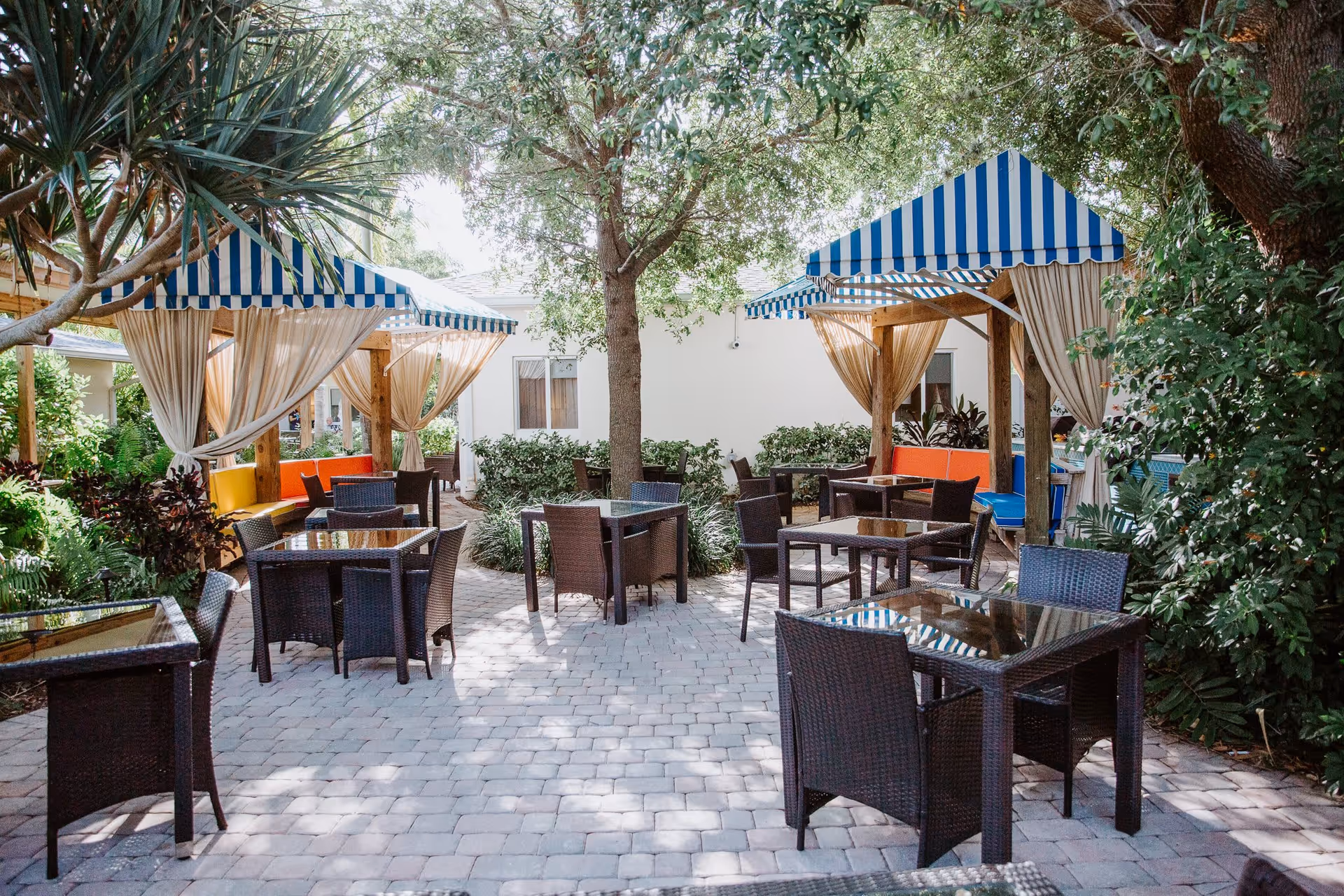 Outdoor patio area with several glass-top tables and wicker chairs arranged on a stone-paved ground. Two wooden pergolas with blue and white striped canopies and beige curtains provide shaded seating with colorful cushions. The area is surrounded by lush green trees and plants, creating a serene and inviting atmosphere.