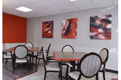 A dining area with several tables and chairs. The walls are painted gray and red, and three abstract red, black, and white paintings hang on the wall. The chairs have a patterned backrest and dark wooden frames.