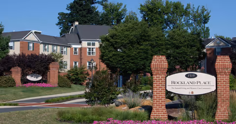 Entrance to Rockland Place, an assisted living and memory care community, featuring a brick sign with the facility name and address 1519, surrounded by landscaped greenery and flowers, with a multi-story brick and beige building in the background under a clear blue sky.