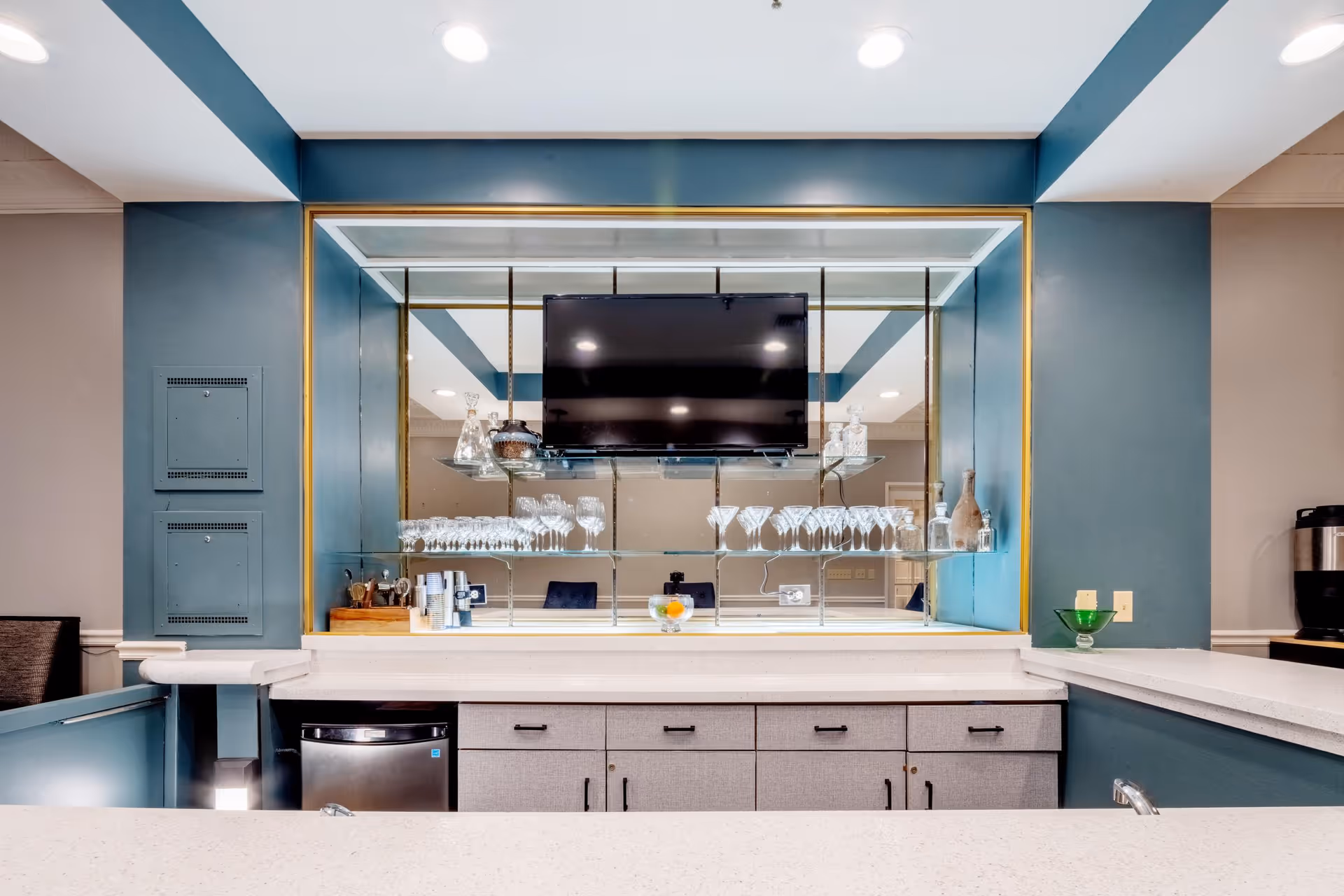A modern bar area with a countertop, hanging wine glasses, a wall-mounted TV, and storage cabinets.