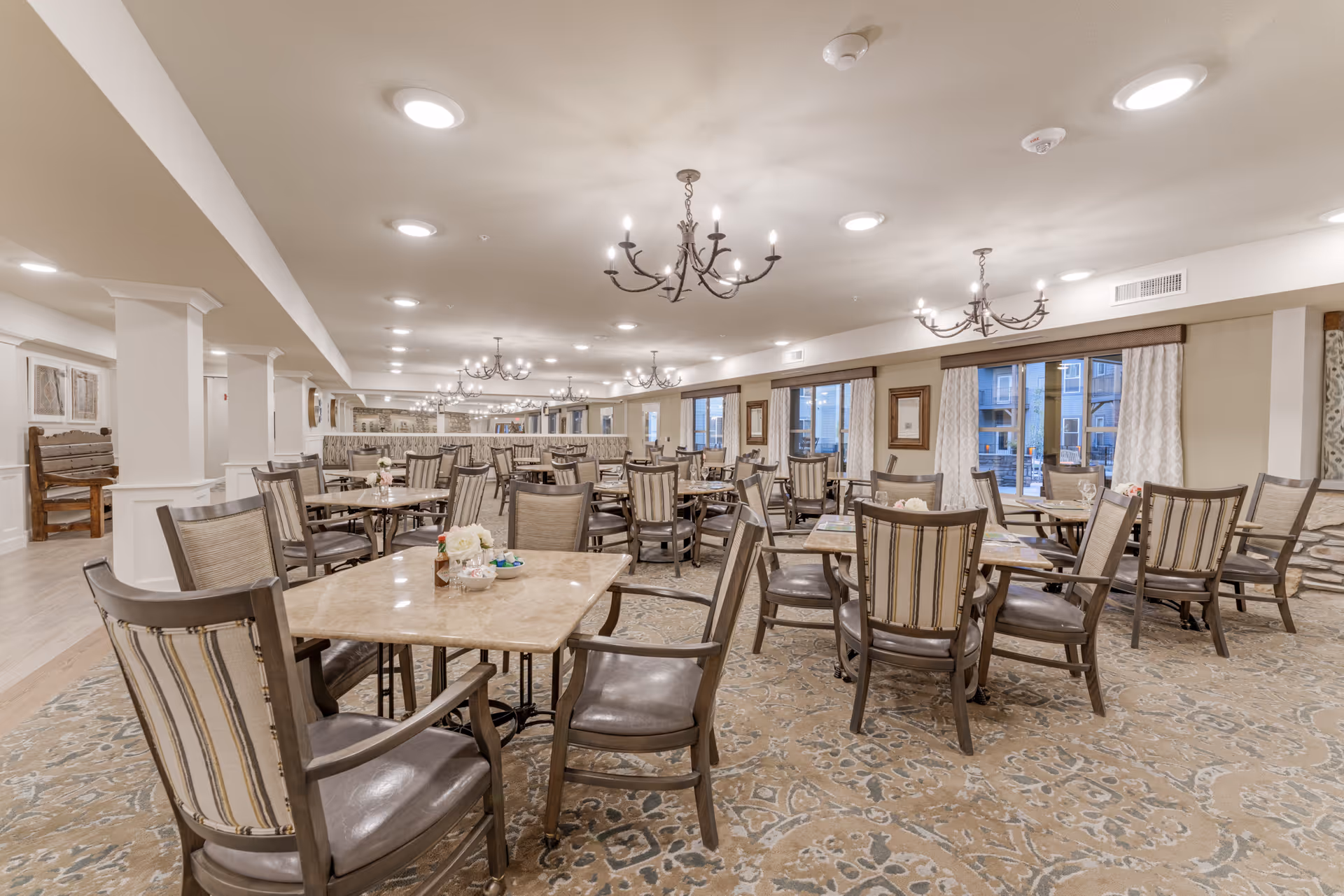 A spacious dining room in a senior living facility with multiple square tables and cushioned chairs arranged neatly. The room features beige walls, large windows with curtains, chandeliers hanging from the ceiling, and a patterned carpet covering the floor. The atmosphere is warm and inviting.
