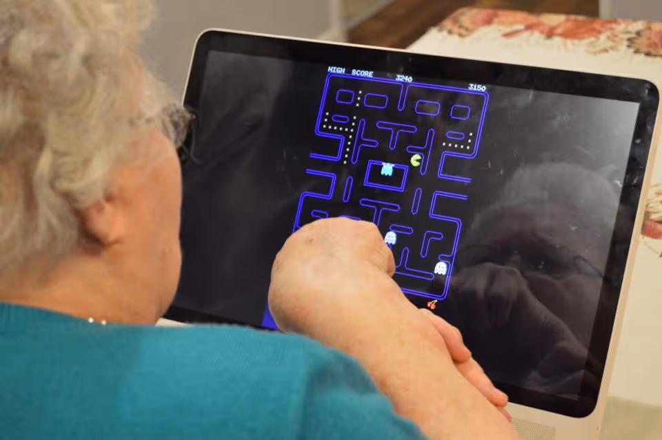 An elderly person with white hair and glasses is playing a classic Pac-Man video game on a touchscreen device, with their hand interacting with the screen.