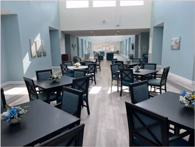 A bright dining room with multiple black tables and blue cushioned chairs arranged neatly. Each table has a small floral centerpiece. The room has light blue walls, large windows near the ceiling allowing natural light, and light wood flooring.