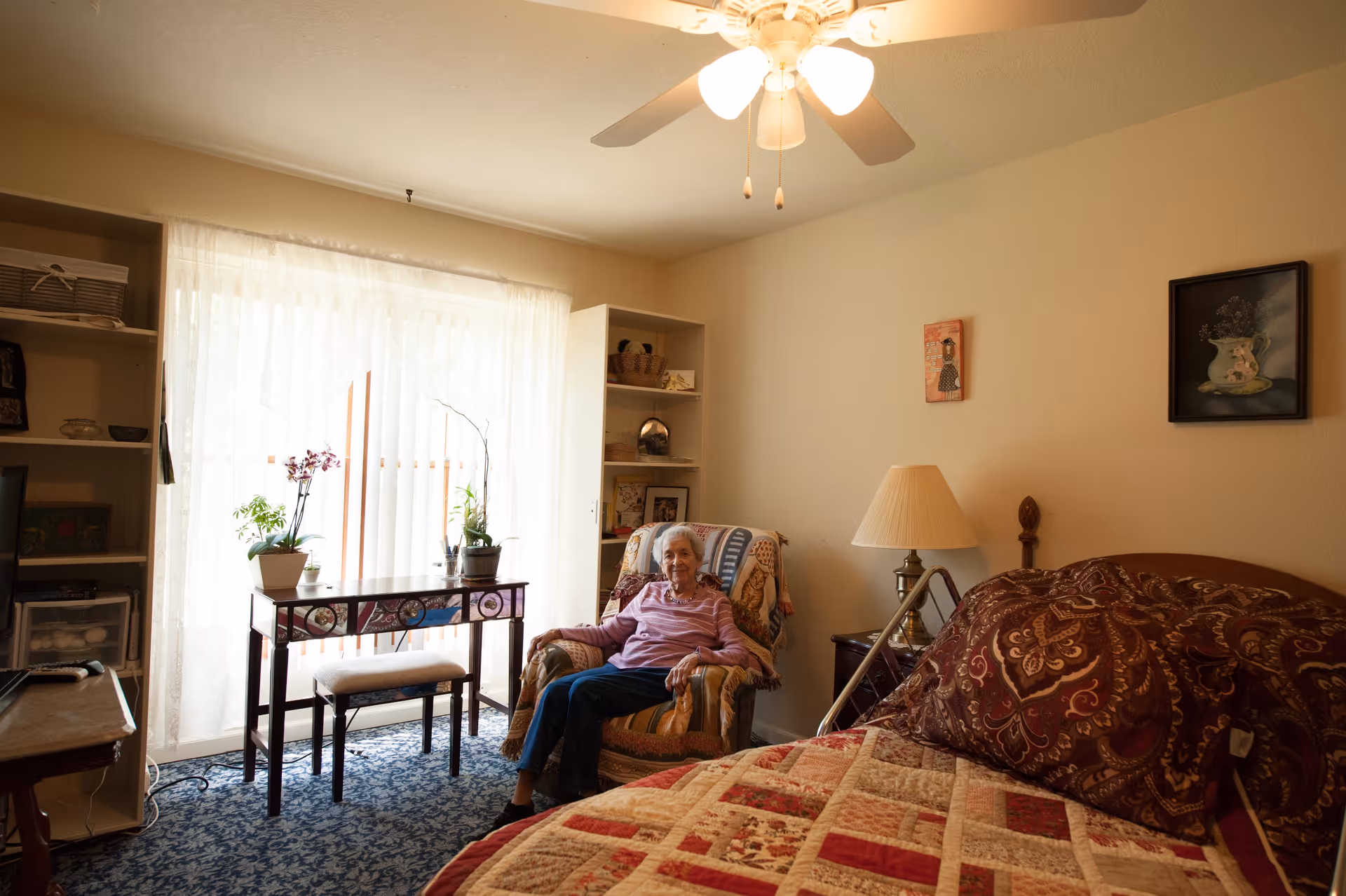 A cozy bedroom with a large bed covered in a patterned quilt and pillows. An elderly woman sits comfortably in an armchair near a window with sheer curtains, next to a small table with potted plants. The room has a ceiling fan with lights, a lamp on a nightstand, and shelves with various items.