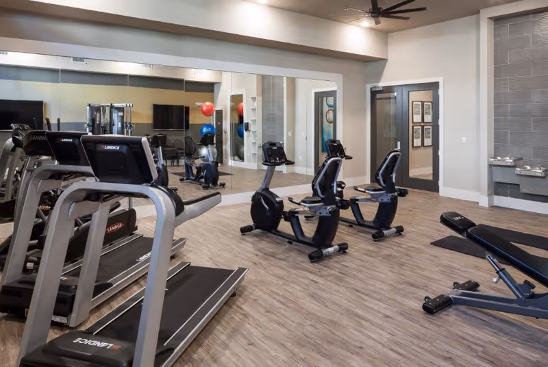 Interior view of a fitness room with exercise equipment including treadmills, stationary bikes, a weight bench, and exercise balls. The room has wood flooring, a large mirror on one wall, and a ceiling fan.