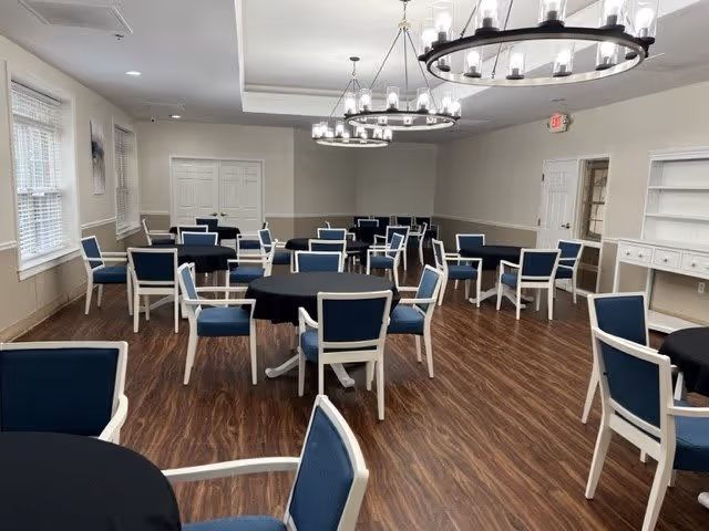 A spacious dining room with multiple round tables covered with black tablecloths and surrounded by blue cushioned chairs with white frames. The room has wood flooring, large windows with blinds, white walls, and two modern circular chandeliers hanging from the ceiling.