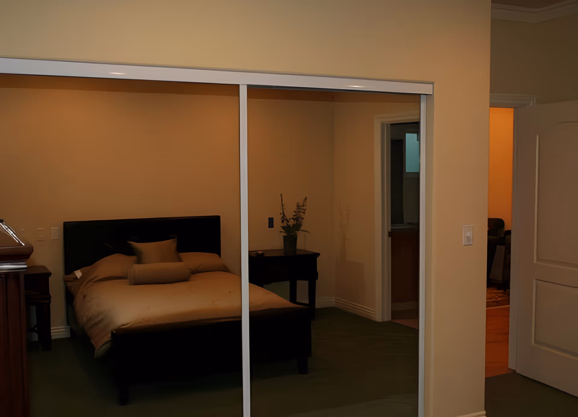 A bedroom with a bed that has beige bedding and pillows. There is a nightstand with a vase of flowers next to the bed. The room is reflected in sliding mirrored closet doors. An open door reveals a glimpse of another room with warm lighting.