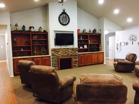 A cozy living room area with four brown upholstered armchairs arranged around a stone fireplace with a mounted flat-screen TV above it. On either side of the fireplace are wooden built-in shelves and cabinets decorated with small birdhouses and other decorative items. The room has a high vaulted ceiling with recessed lighting and a large wall clock above the TV.