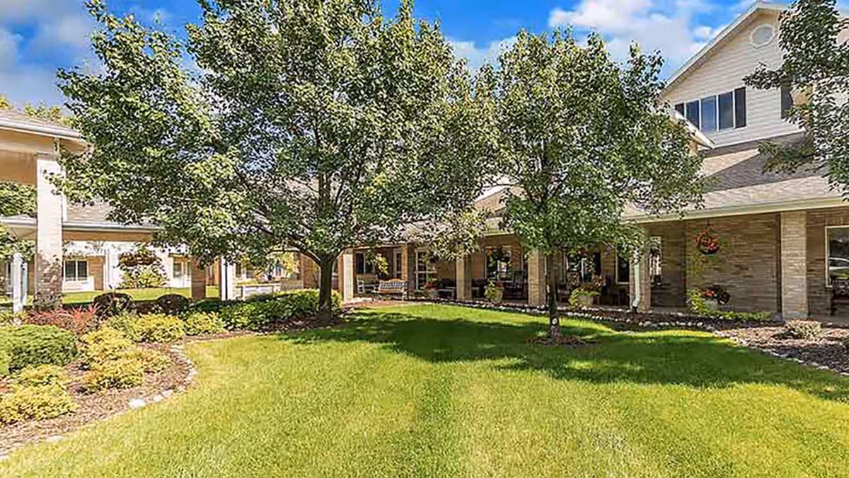 A sunny outdoor garden area at Holiday Genesee Gardens with green grass, two leafy trees, and a building with a covered porch and hanging flower baskets in the background.