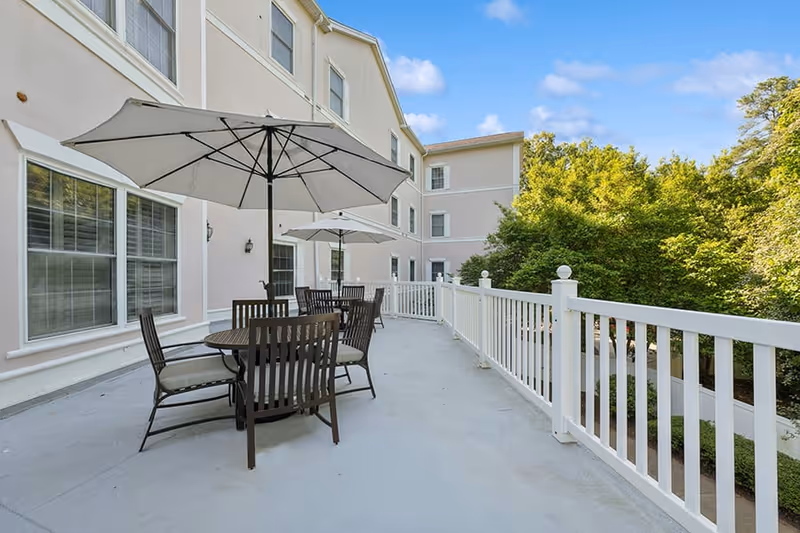 Outdoor patio area at HeartFields at Cary with round tables and chairs under large white umbrellas, adjacent to a light-colored building with multiple windows, surrounded by green trees under a blue sky with some clouds.