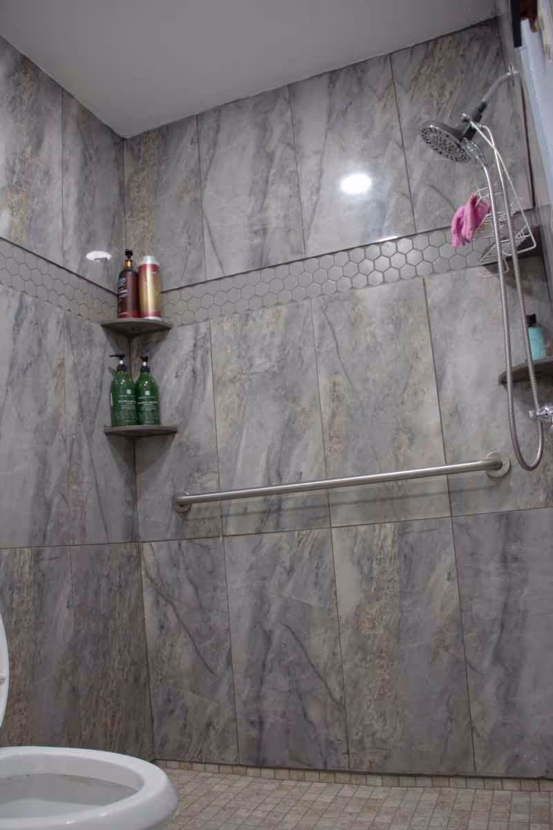 Shower area with gray marble-like tiled walls and a strip of hexagonal tiles. There are two corner shelves holding bottles of shampoo and soap. A silver grab bar is mounted on the wall, and a handheld showerhead is attached to the right wall with a pink washcloth hanging on the shower caddy. The edge of a white toilet seat is visible at the bottom left.