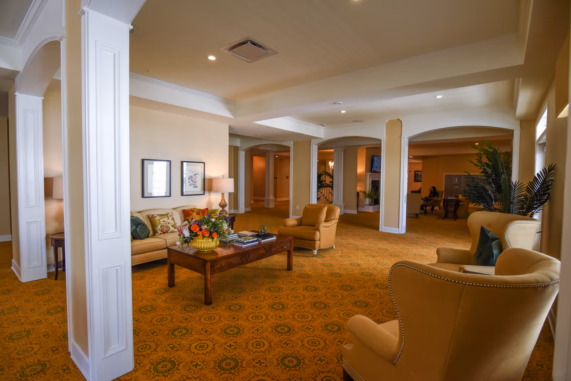 A spacious and well-lit living room area with beige walls and patterned carpet. The room features comfortable seating including a sofa, armchairs, and a wooden coffee table with a floral arrangement and books. Decorative columns and arches separate different sections of the room, and there are framed pictures on the wall and potted plants adding to the cozy atmosphere.