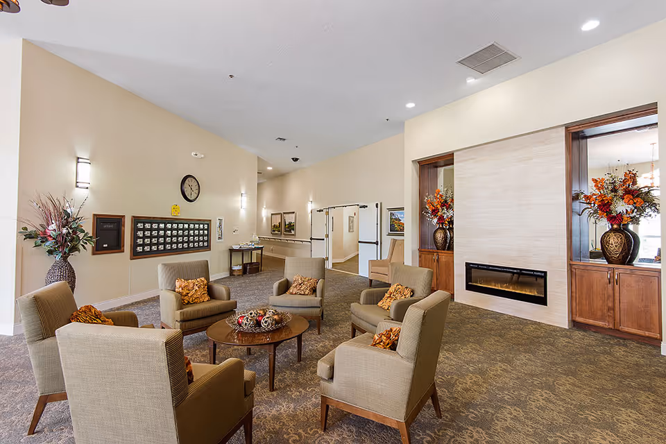 A cozy assisted living facility common area with six beige armchairs arranged around a round wooden coffee table. The room features a modern electric fireplace set in a tiled wall with wooden cabinets on either side, each holding a large vase with colorful floral arrangements. The walls are painted light beige, and there is a clock and a mail organizer on one wall. The carpet is patterned in neutral tones, and the space is softly lit with wall sconces and recessed ceiling lights.