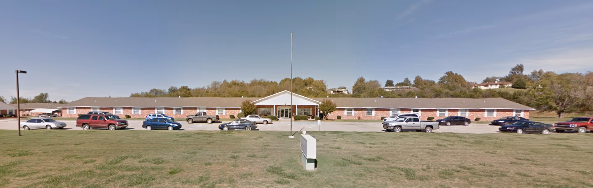 Wide exterior view of a single-story brick building with a peaked entrance, several parked cars in front, and a large grassy area in the foreground under a clear blue sky.