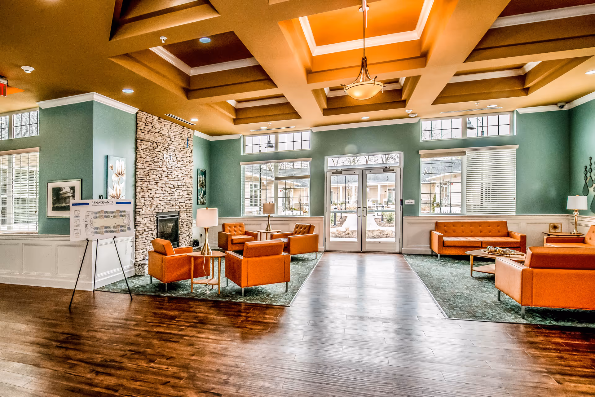 A spacious and well-lit living room area with green walls and a coffered ceiling painted in warm tones. The room features a stone fireplace, several orange leather armchairs and sofas arranged around small tables, large windows letting in natural light, and double glass doors leading outside. There is a floor plan displayed on an easel near the fireplace.
