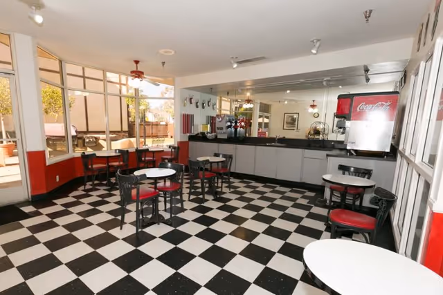 Small cafeteria-style dining area with a black-and-white checkered floor, round tables with red-cushioned chairs, a drink station, and large windows.