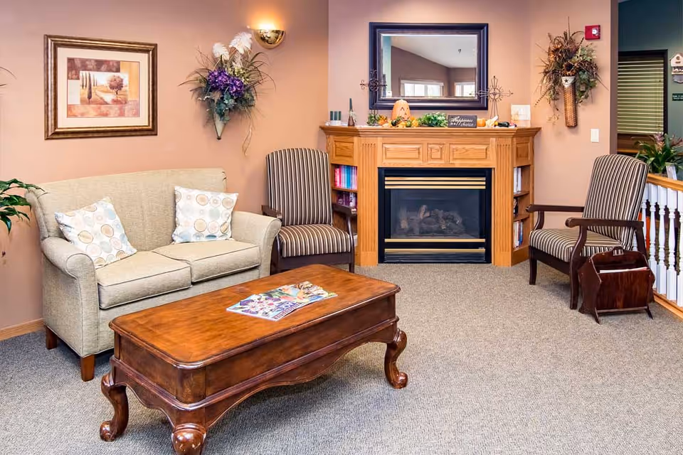 A cozy living room area with a beige couch adorned with two patterned pillows, two striped armchairs, a wooden coffee table with magazines on top, and a wooden fireplace with a mirror above it. The walls are decorated with a framed picture and floral arrangements.