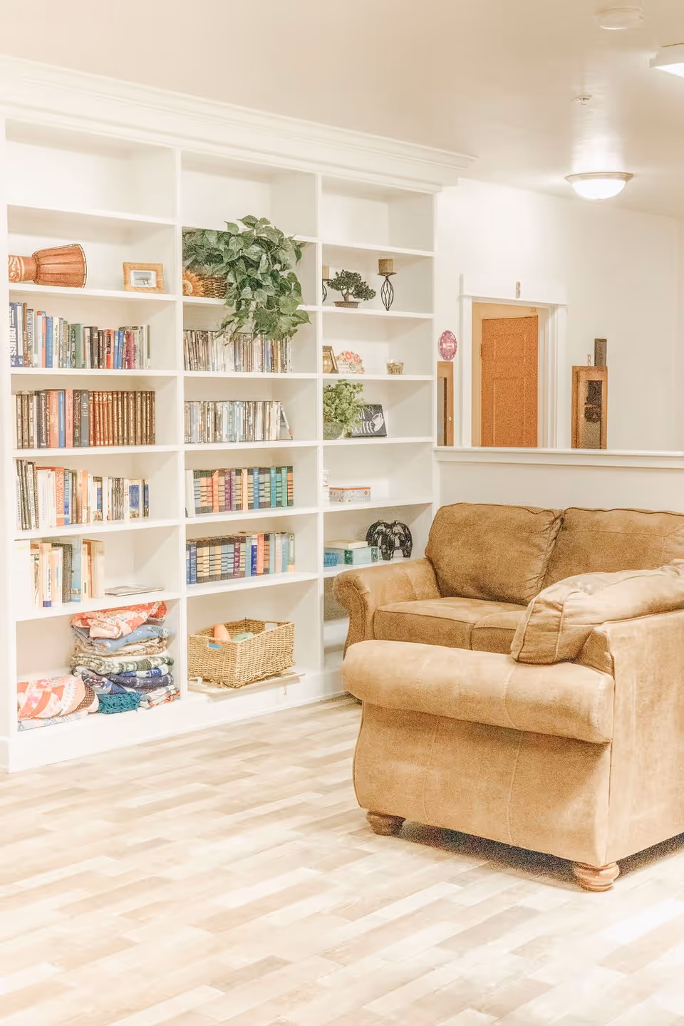 A cozy living room area with a brown leather armchair and a large white built-in bookshelf filled with books, plants, and decorative items. The room has light-colored wooden flooring and white walls with a doorway in the background.