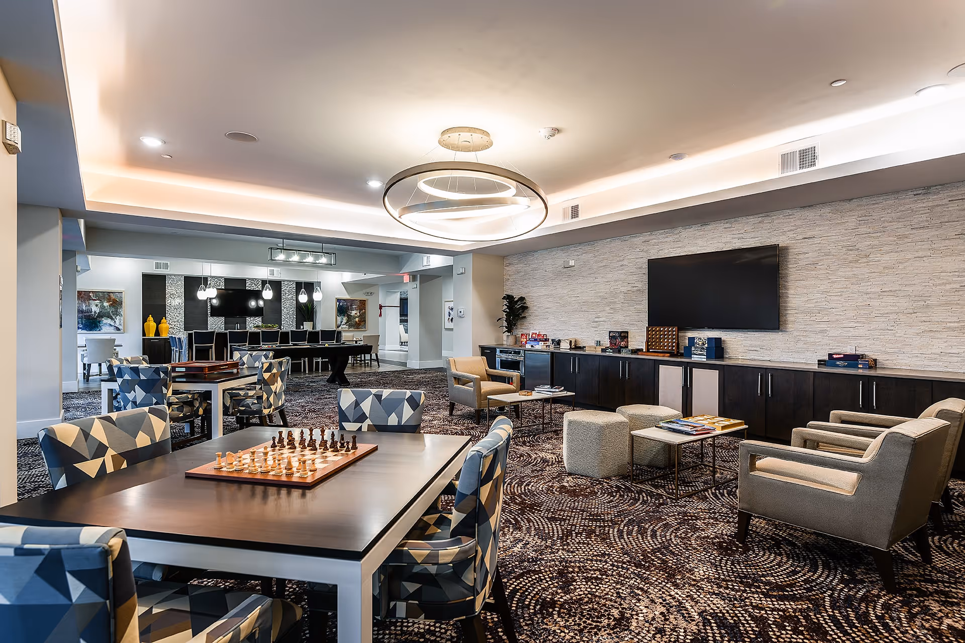 A spacious and modern common area in a senior living facility featuring a chess table with patterned chairs in the foreground, comfortable armchairs, ottomans, and a large flat-screen TV mounted on a textured stone wall. The room is well-lit with a contemporary circular chandelier and recessed lighting, and there are additional tables and chairs in the background near decorative artwork and pendant lights.