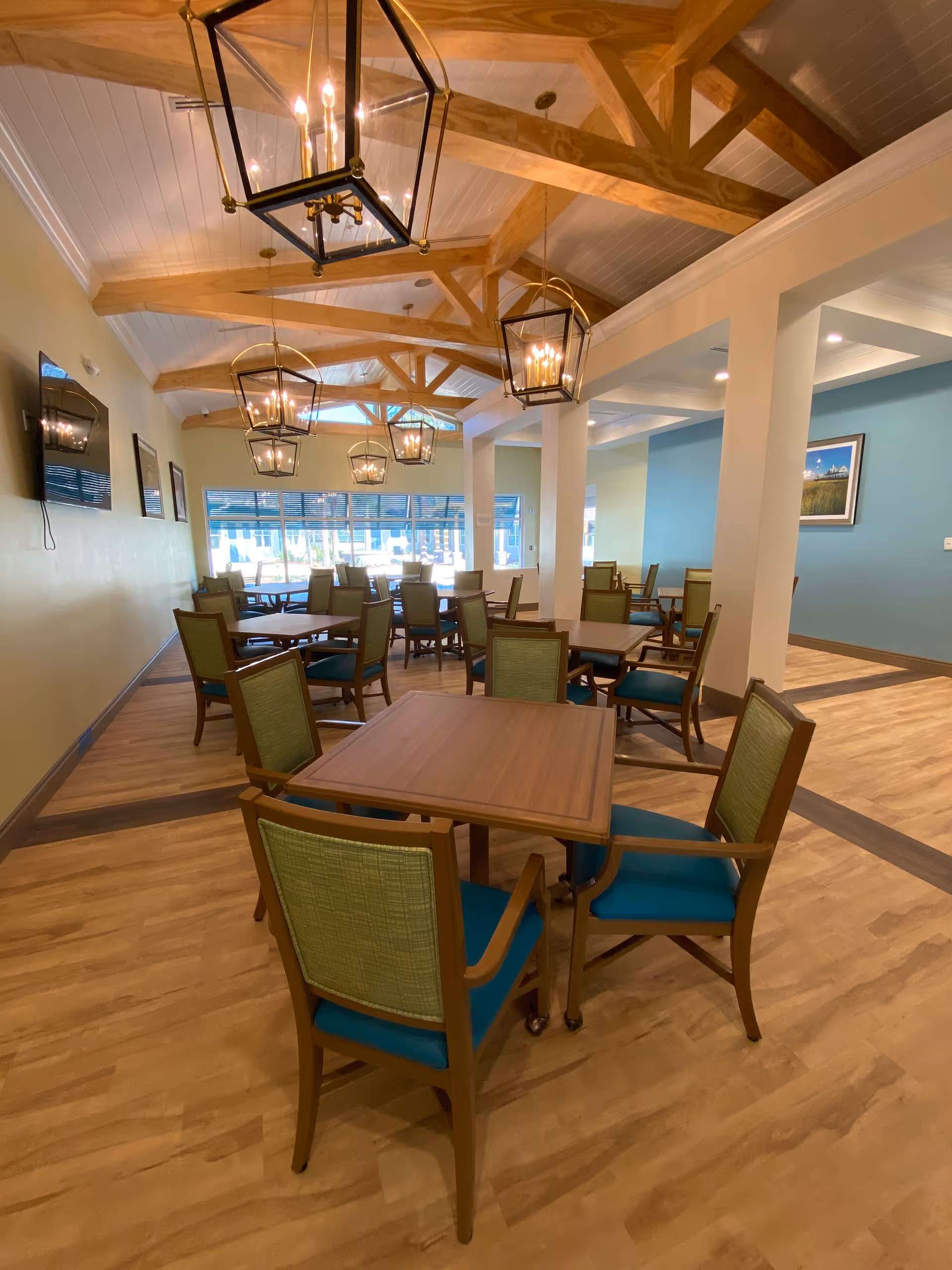 A spacious dining room with multiple wooden tables and green cushioned chairs. The room features wooden beams on the ceiling with hanging lantern-style light fixtures. Large windows at the far end allow natural light to fill the space, and the walls are painted in soft yellow and blue tones.