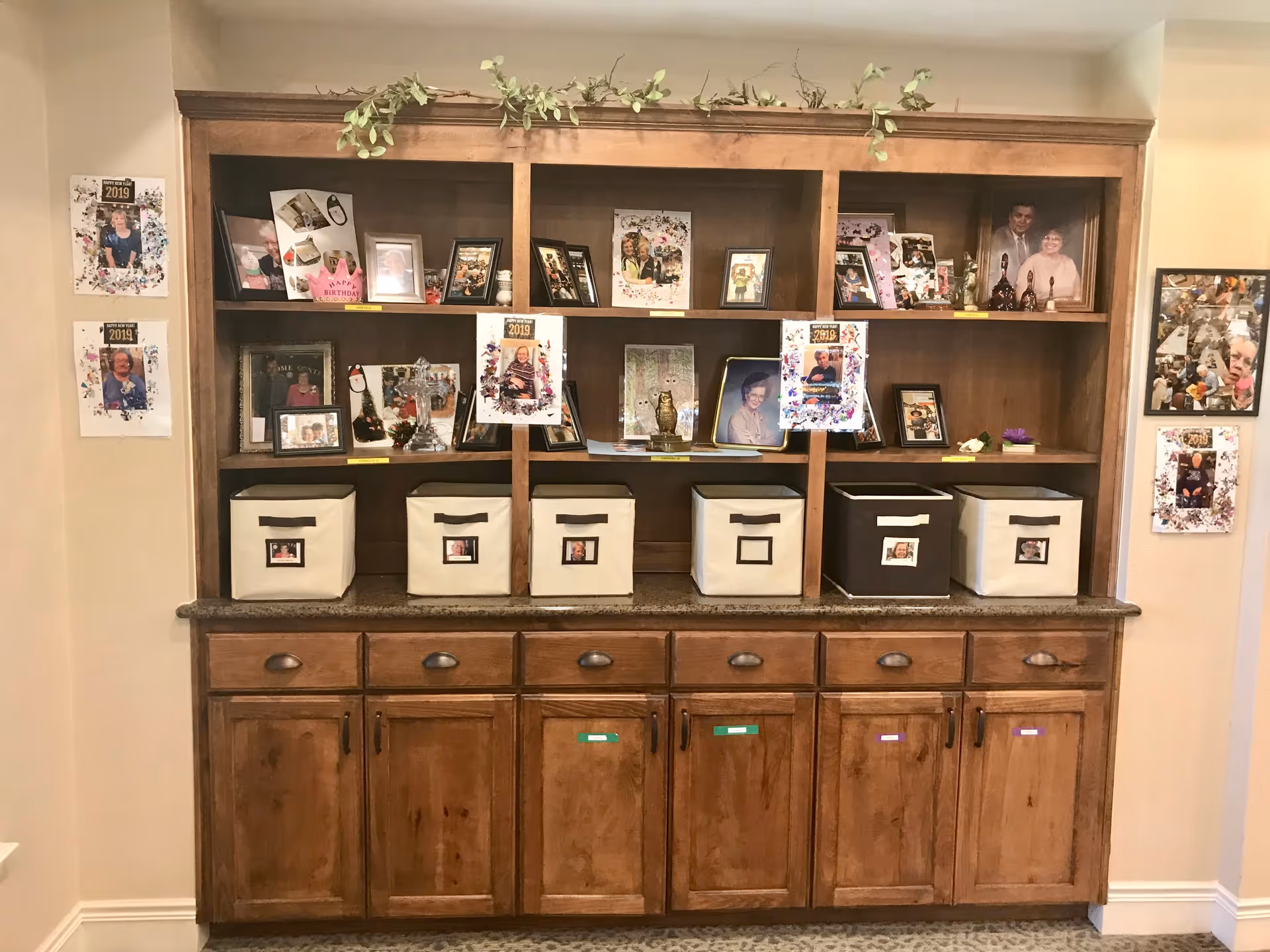 Wooden built-in shelving and lower cabinets displaying framed photos, decorative items, and labeled storage bins in a communal interior space.