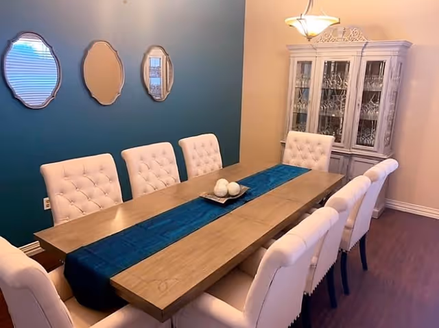 A dining room with a long wooden table covered by a blue table runner and decorated with a centerpiece of three white decorative balls. The table is surrounded by eight white tufted chairs. On the wall behind the table, there are three decorative mirrors with ornate frames. A white china cabinet filled with glassware is positioned against the adjacent wall, and a hanging light fixture illuminates the room.