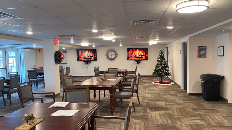 Interior view of a community room with multiple wooden tables and chairs arranged neatly. Two flat-screen TVs on the far wall display a virtual fireplace. A decorated Christmas tree stands to the right side of the room. The room has beige walls, a patterned carpet, and ceiling lights. There are wreaths hanging on the walls and a trash bin near the right wall.