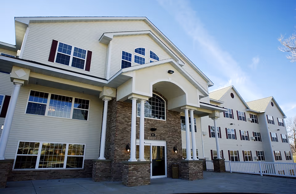Exterior front view of a multi-story senior living facility building with beige siding, brick accents, multiple windows with red shutters, and a covered entrance supported by white columns under a clear blue sky.
