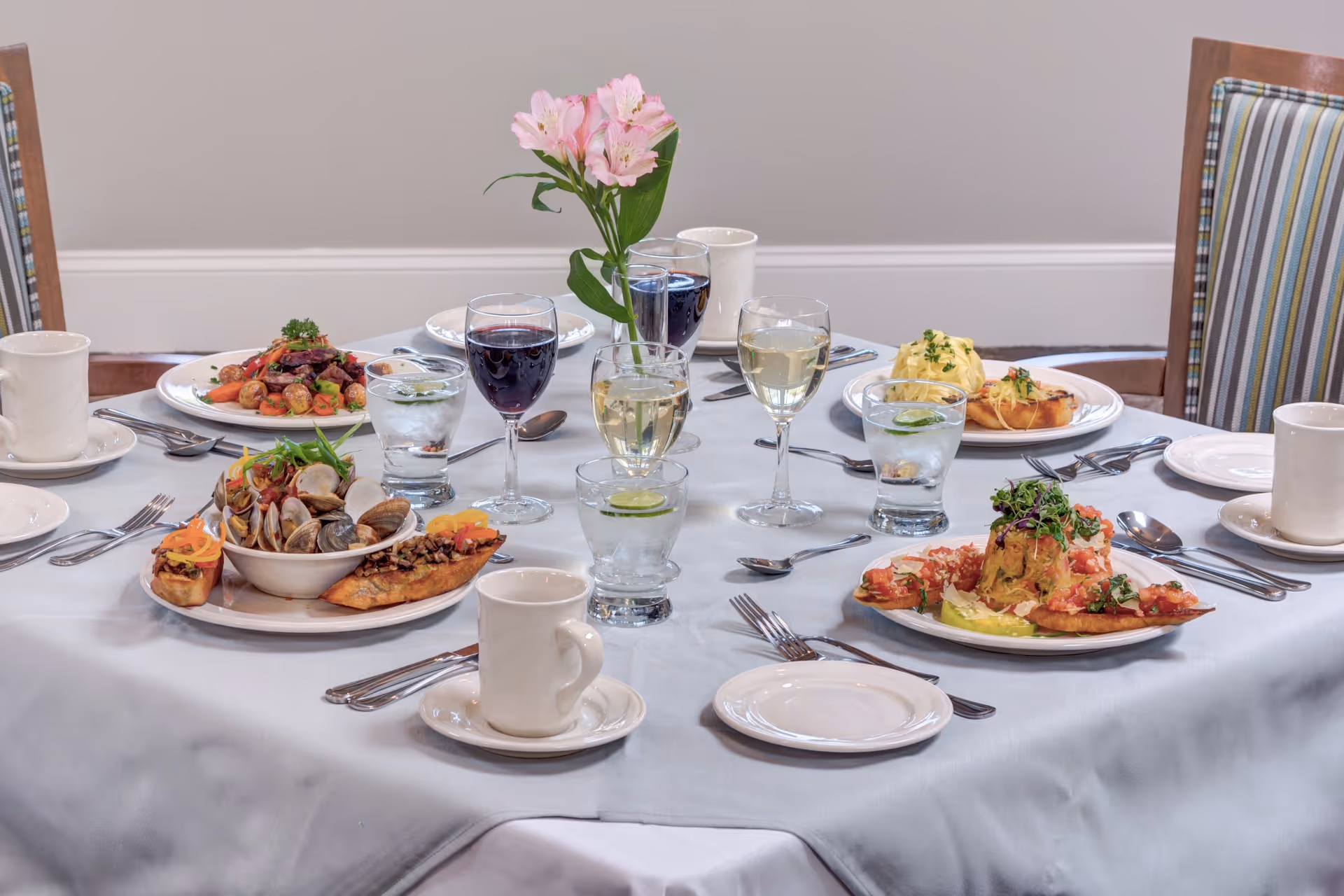 A dining table set with plates of gourmet food, glasses of water with lime, wine glasses filled with red and white wine, white coffee cups, and silverware. A small vase with pink flowers is placed in the center of the table. The chairs around the table have striped upholstery.