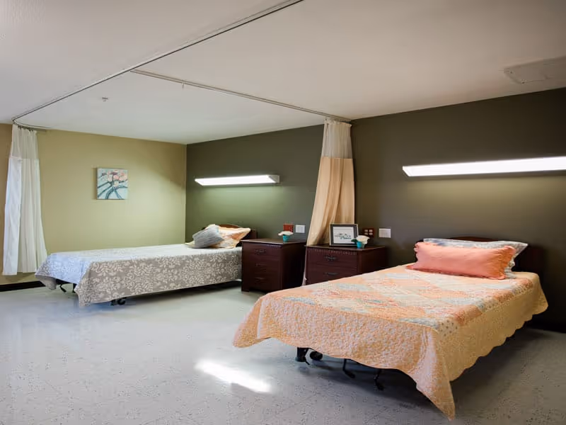 Two single beds in a shared nursing/rehab room with nightstands and privacy curtains.