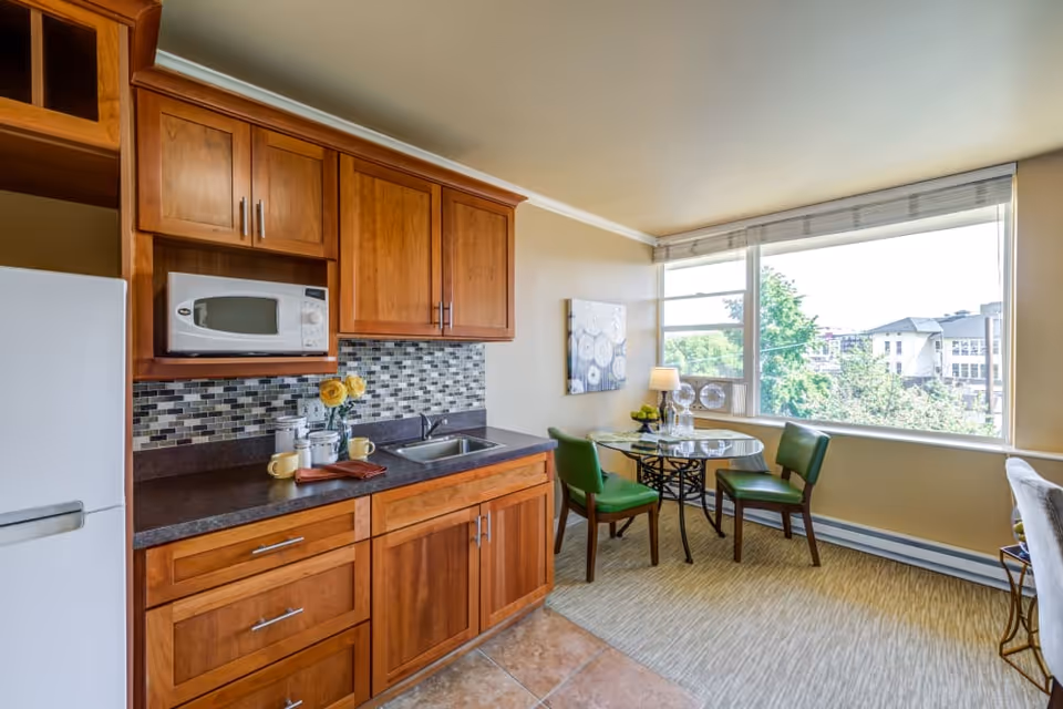 A bright kitchen and dining area with wooden cabinets, a microwave, a sink, and a countertop with cups and a vase of yellow flowers. Next to the kitchen is a small round glass dining table with three green chairs, a table lamp, and a large window showing trees and buildings outside.