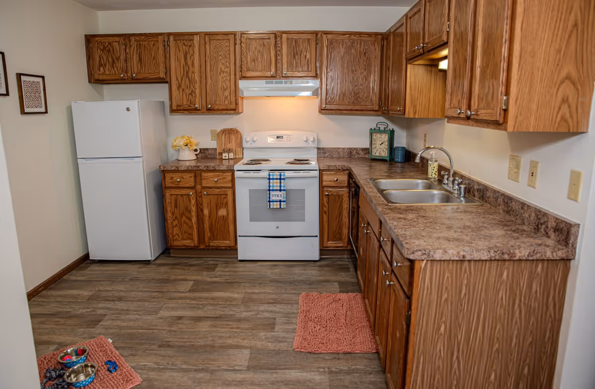 A kitchen with wooden cabinets, a white refrigerator, a white electric stove with a blue and white checkered towel hanging on the handle, a double sink, and a brown countertop. The floor is wood-patterned, and there are two pink rugs on the floor. On the countertop, there is a decorative clock, a small container, and a vase with yellow flowers.