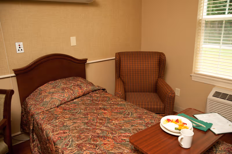 Single-bed patient room with a patterned bedspread, armchair, window, and a meal tray on a small table.