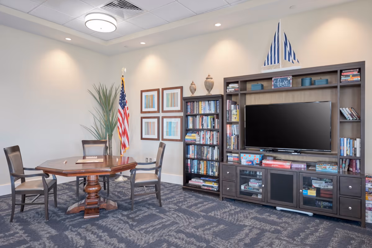 A cozy common area with a wooden octagonal table surrounded by four chairs, a tall bookshelf filled with books, a large entertainment center with a flat-screen TV, board games, and decorative items including a model sailboat and two vases. An American flag and a tall plant are positioned in the corner, with four framed abstract artworks on the wall.