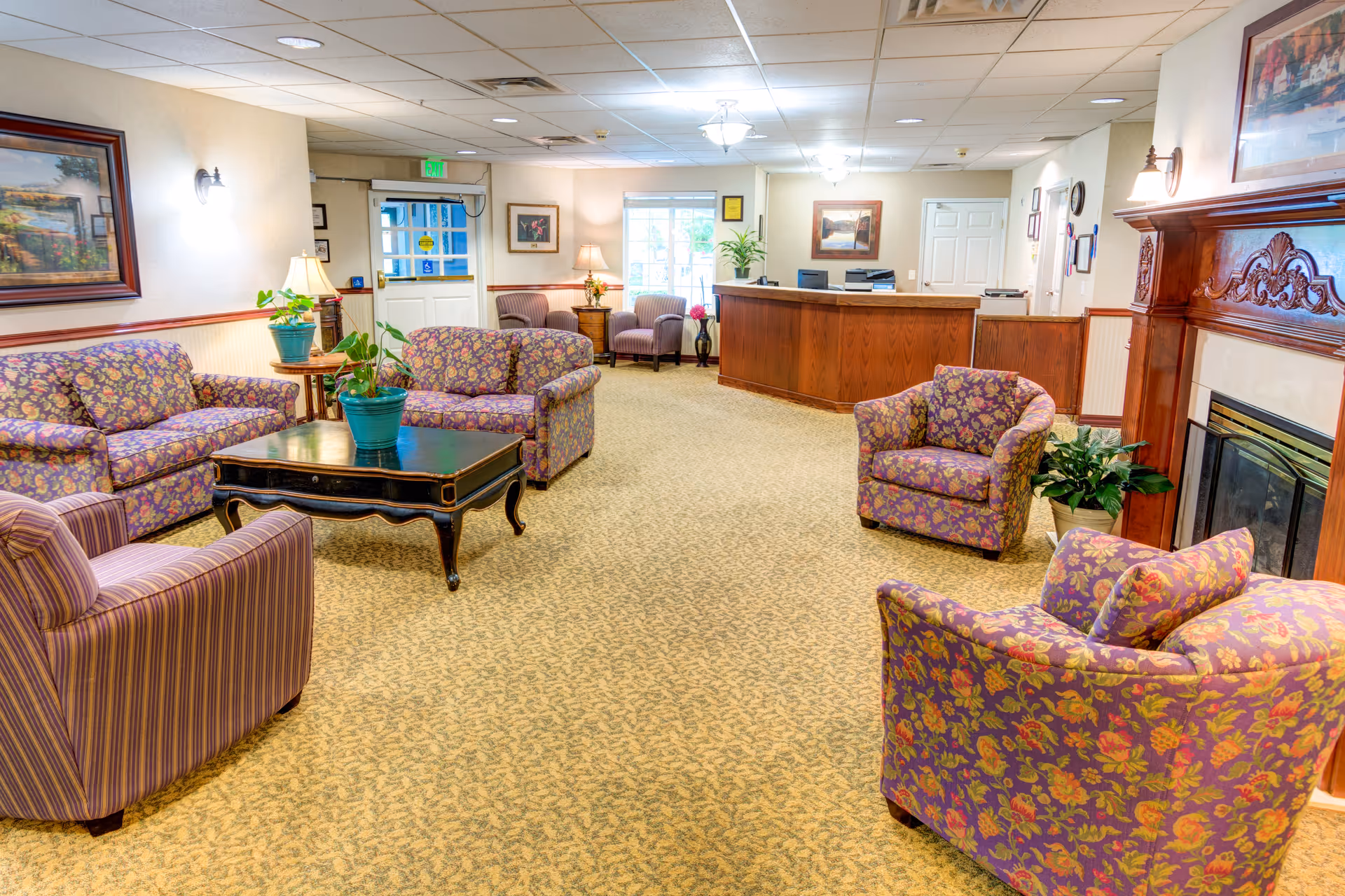 A cozy senior living facility lounge area with floral patterned sofas and armchairs arranged around a dark wooden coffee table. There is a fireplace with a decorative wooden mantle on the right side, potted plants on tables and the floor, framed artwork on the walls, and a reception desk in the background near the entrance door.