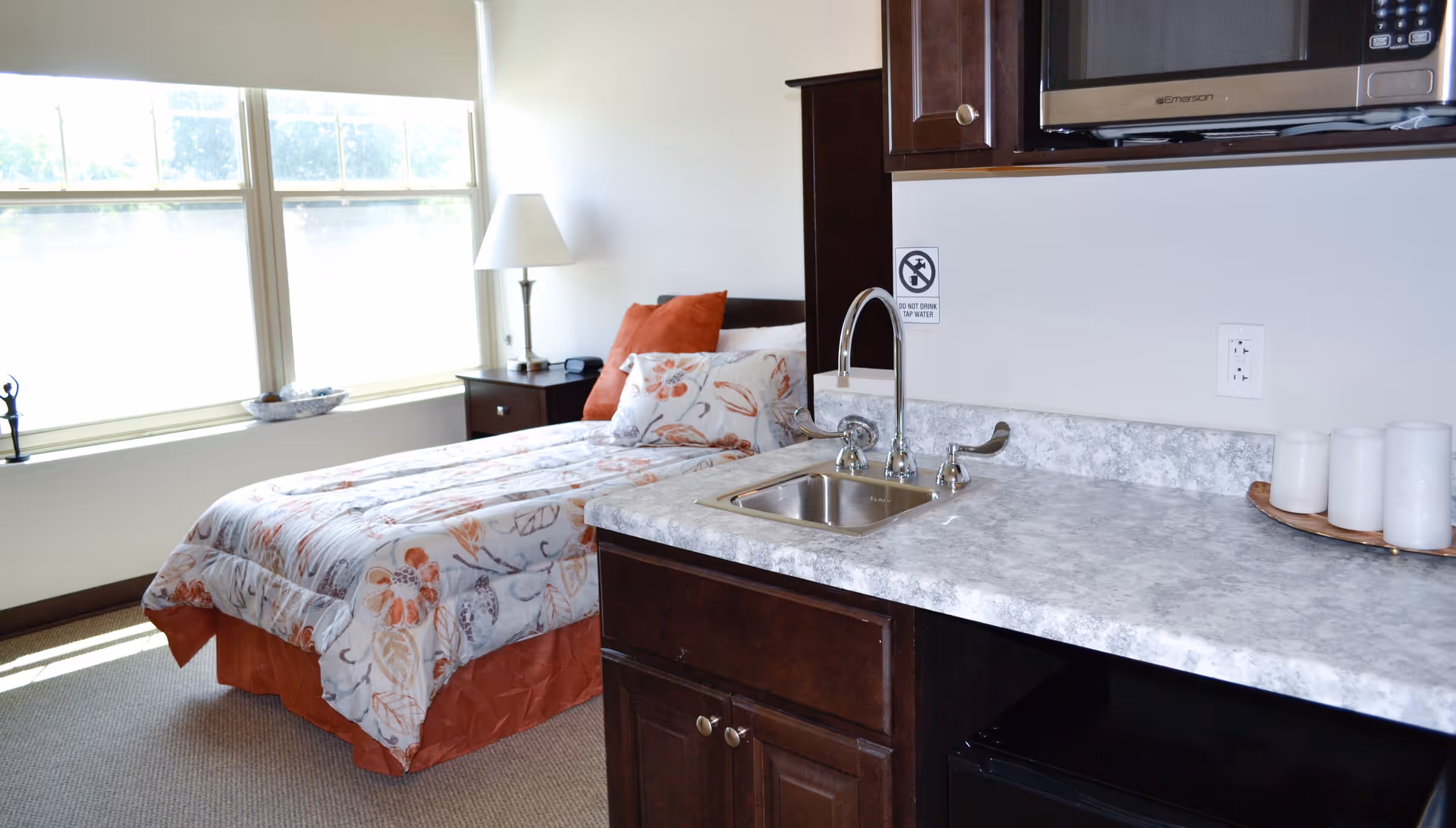 Bright studio bedroom with a bedside table and lamp next to a single bed, and a small kitchenette countertop with sink and microwave.