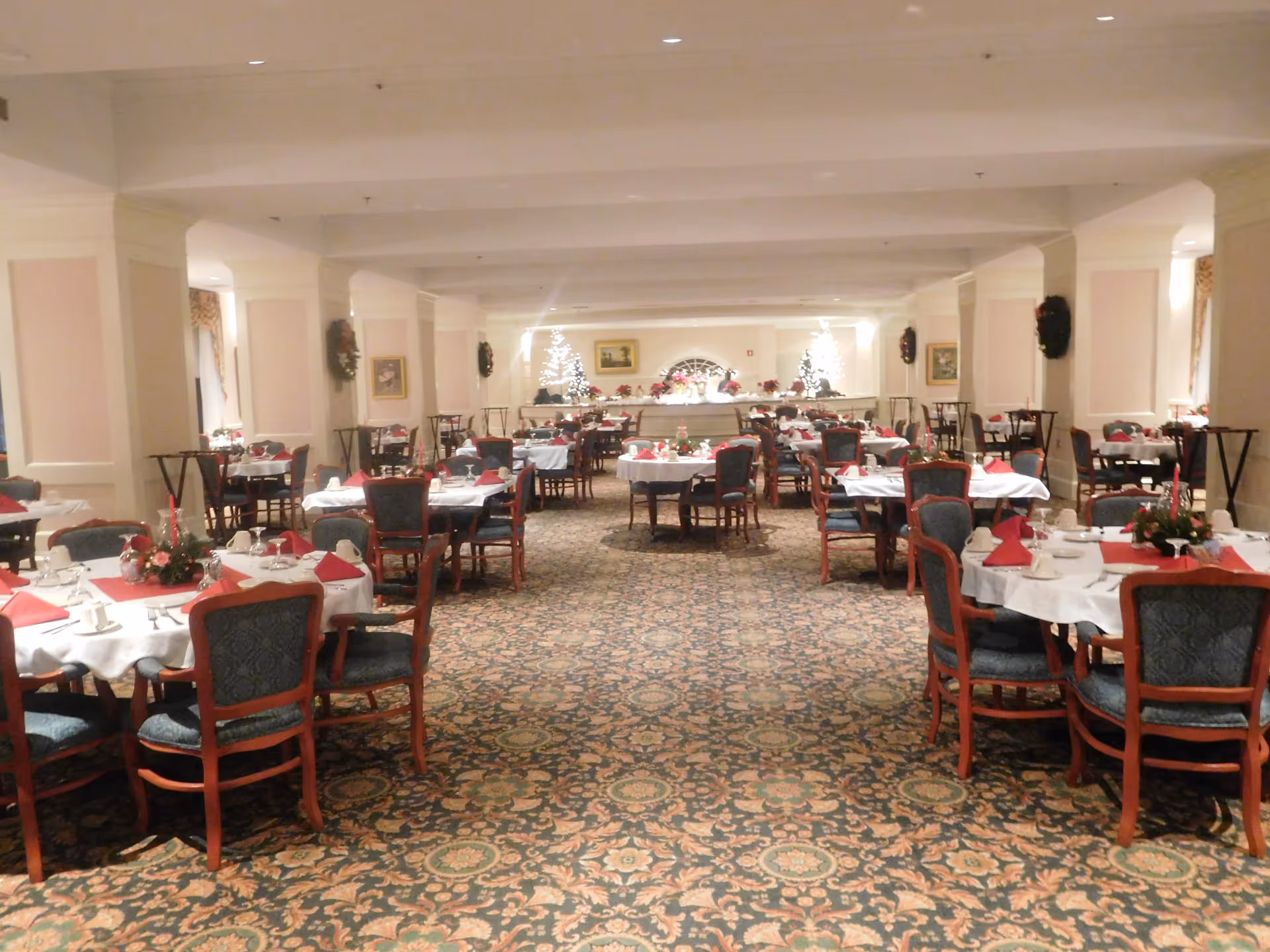 A large dining room with multiple round tables covered with white tablecloths and red napkins. Each table is set with cups, plates, and floral centerpieces. The room is decorated with Christmas wreaths on the walls and two lit Christmas trees at the far end. The carpet has an ornate floral pattern, and the chairs have wooden frames with dark upholstered seats and backs.