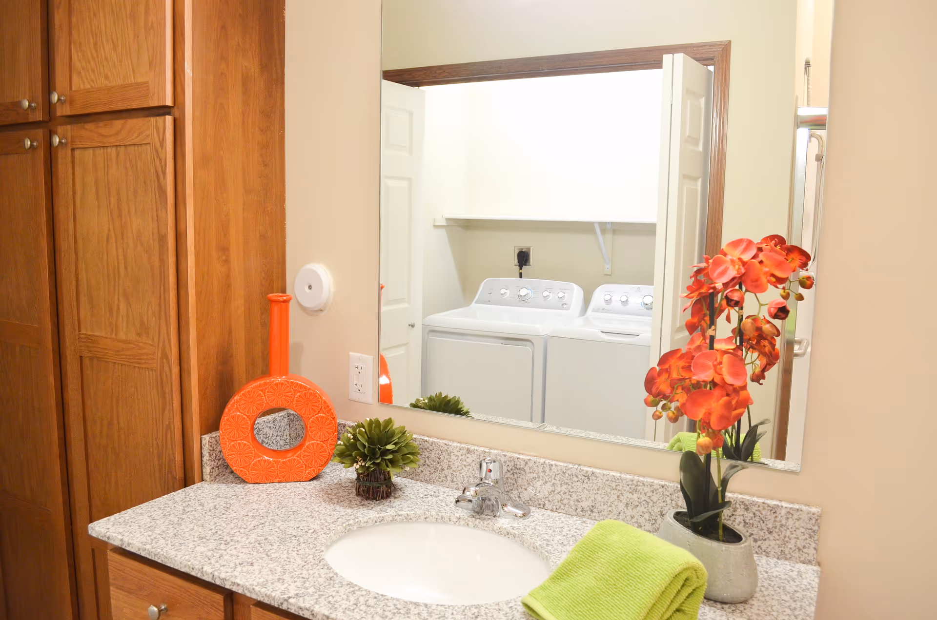 Bathroom vanity with a granite countertop, a sink, a green towel, an orange decorative vase, and a small green plant. A large mirror above the sink reflects a laundry area with a washer and dryer.