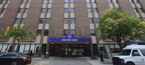 Front view of a tall brick senior care building with a purple entrance sign and trees.