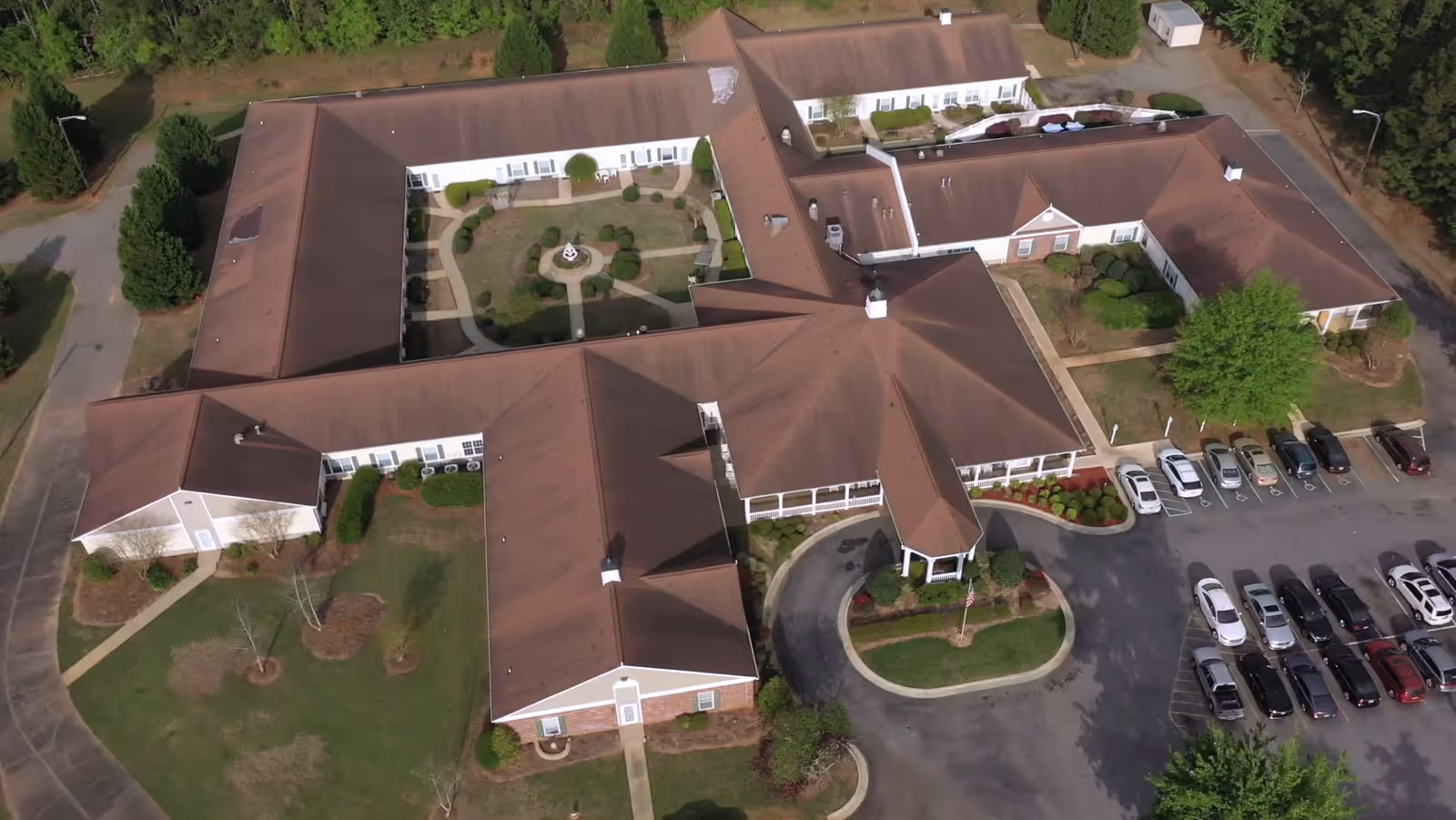 Aerial view of a senior living facility building with a central courtyard garden, surrounded by parking spaces with multiple cars parked. The building has a brown roof and white exterior walls, with pathways and landscaped greenery around it.