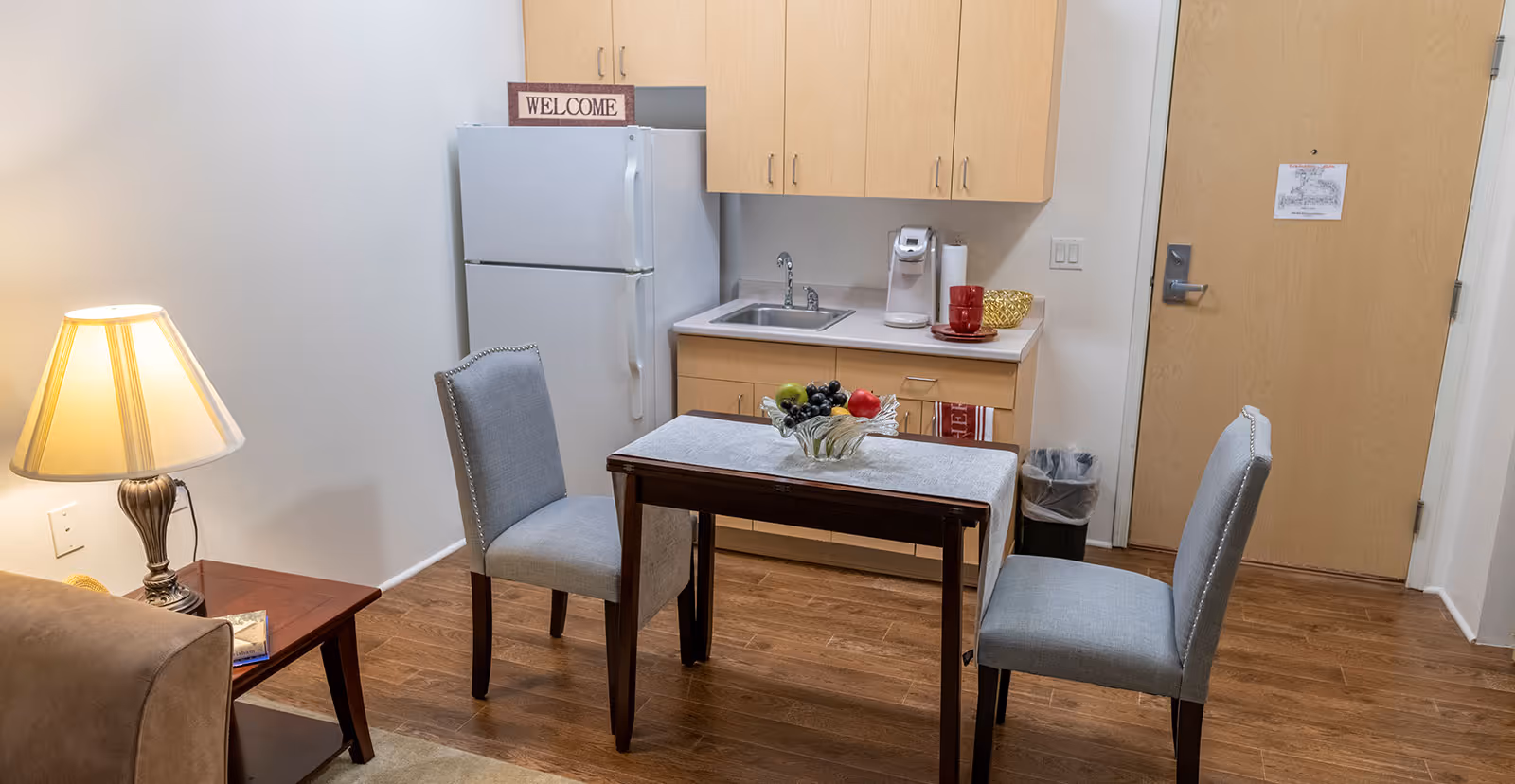 A small kitchen area in a senior living facility with light wood cabinets, a white refrigerator, a sink, and a coffee maker on the counter. In front of the kitchen is a small wooden table with a gray runner and a glass bowl of fruit. Two gray upholstered chairs are placed on either side of the table. To the left, there is a side table with a lamp and a book, and a beige couch is partially visible. The entrance door is visible on the right side of the image.