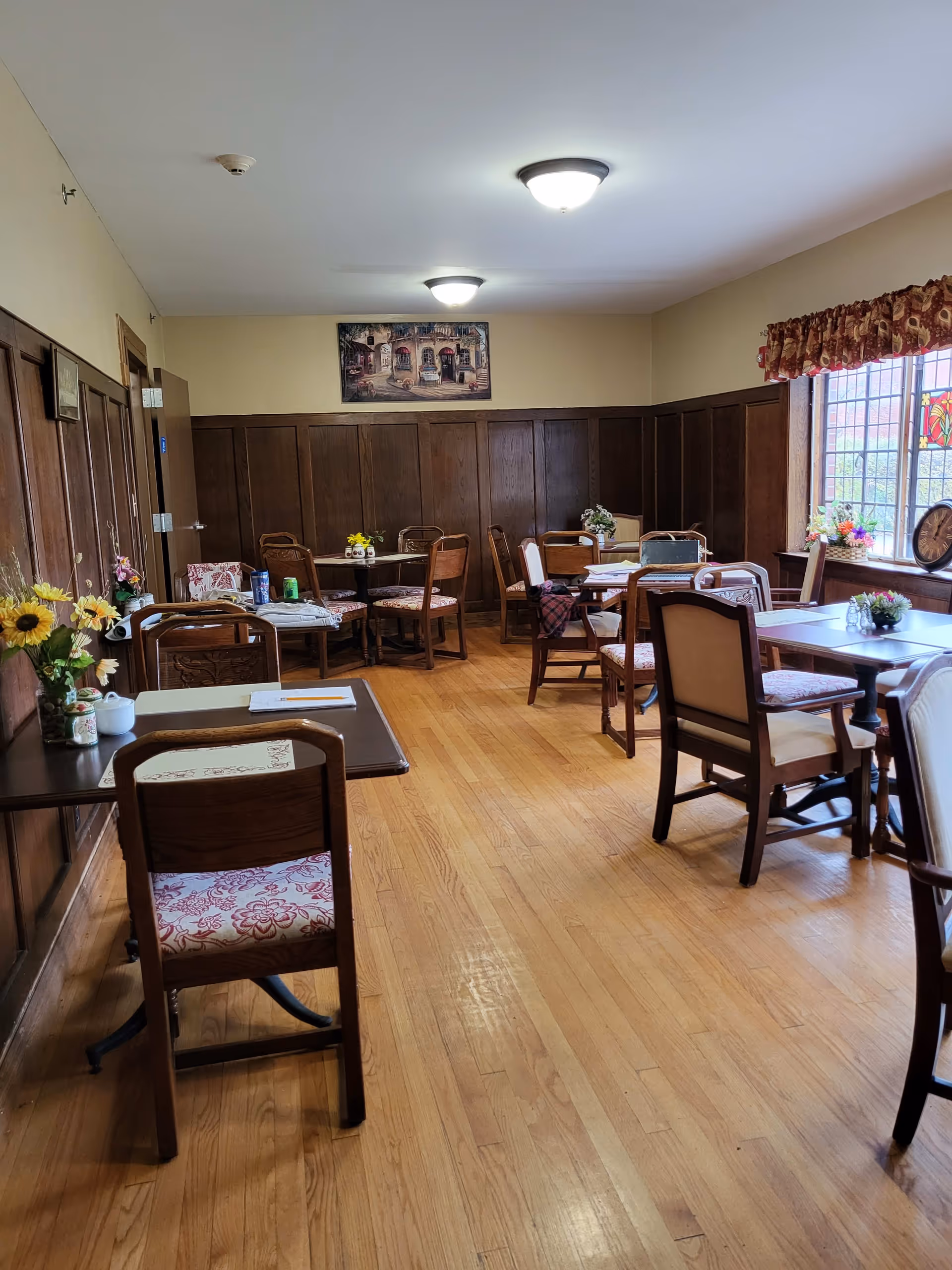A cozy dining room with wooden floors and paneled walls. Several wooden tables and chairs with floral cushions are arranged around the room. The room is decorated with flowers on the tables and a painting on the far wall. A large window with a decorative valance lets in natural light.