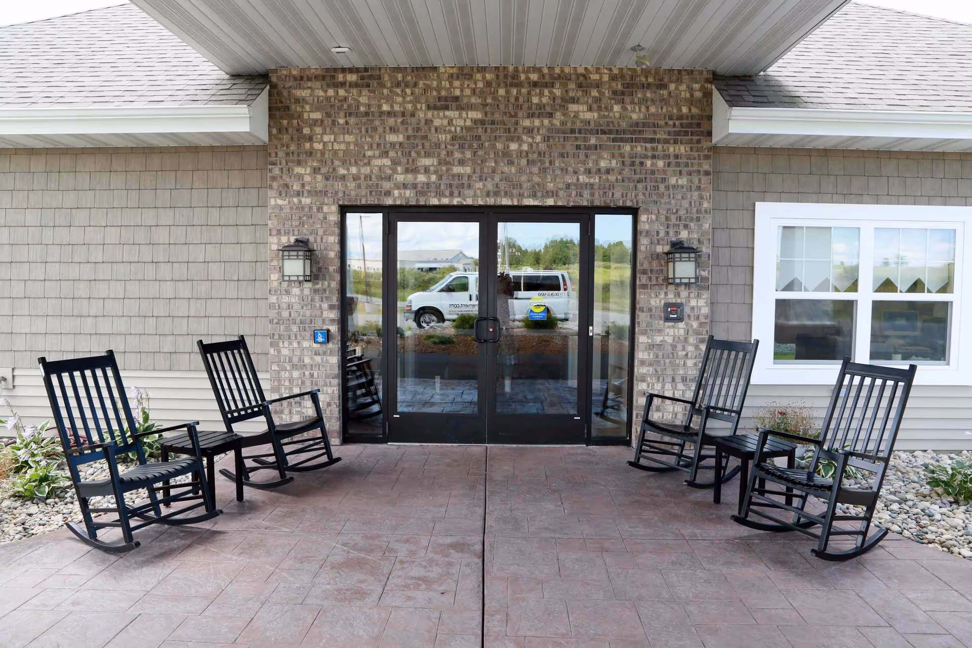 Covered front entrance with six black rocking chairs arranged on a stamped concrete porch in front of glass double doors.
