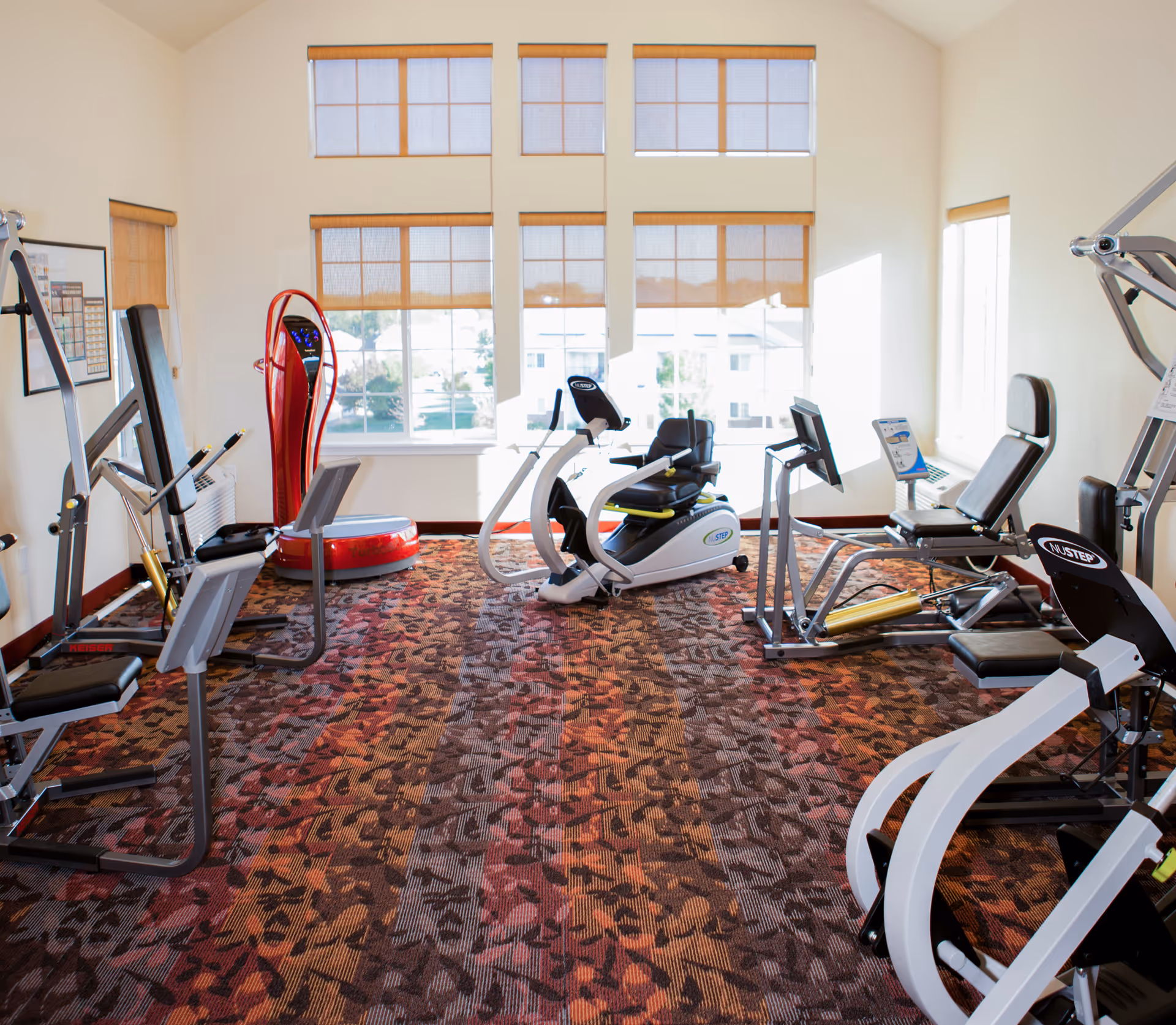 Small fitness room with several exercise machines, large multi-pane windows, and patterned carpet.