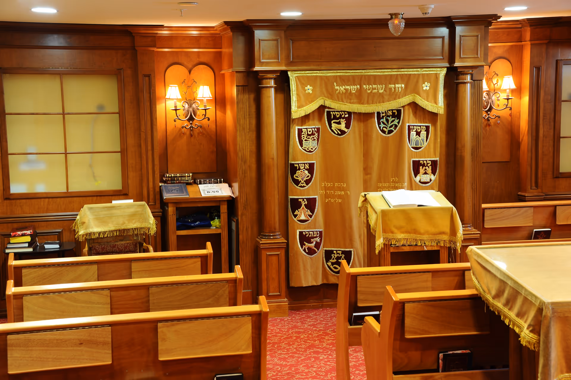 Interior view of a small synagogue or prayer room with wooden pews, a red carpet, and a wooden ark covered with a gold curtain featuring Hebrew text and symbols. The room is warmly lit with wall sconces and has lecterns covered with gold cloths.