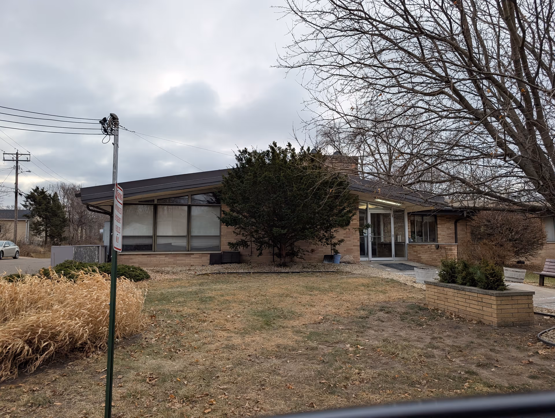 Exterior view of a single-story brick building with large windows and a glass entrance door, surrounded by leafless trees and dry grass under a cloudy sky.