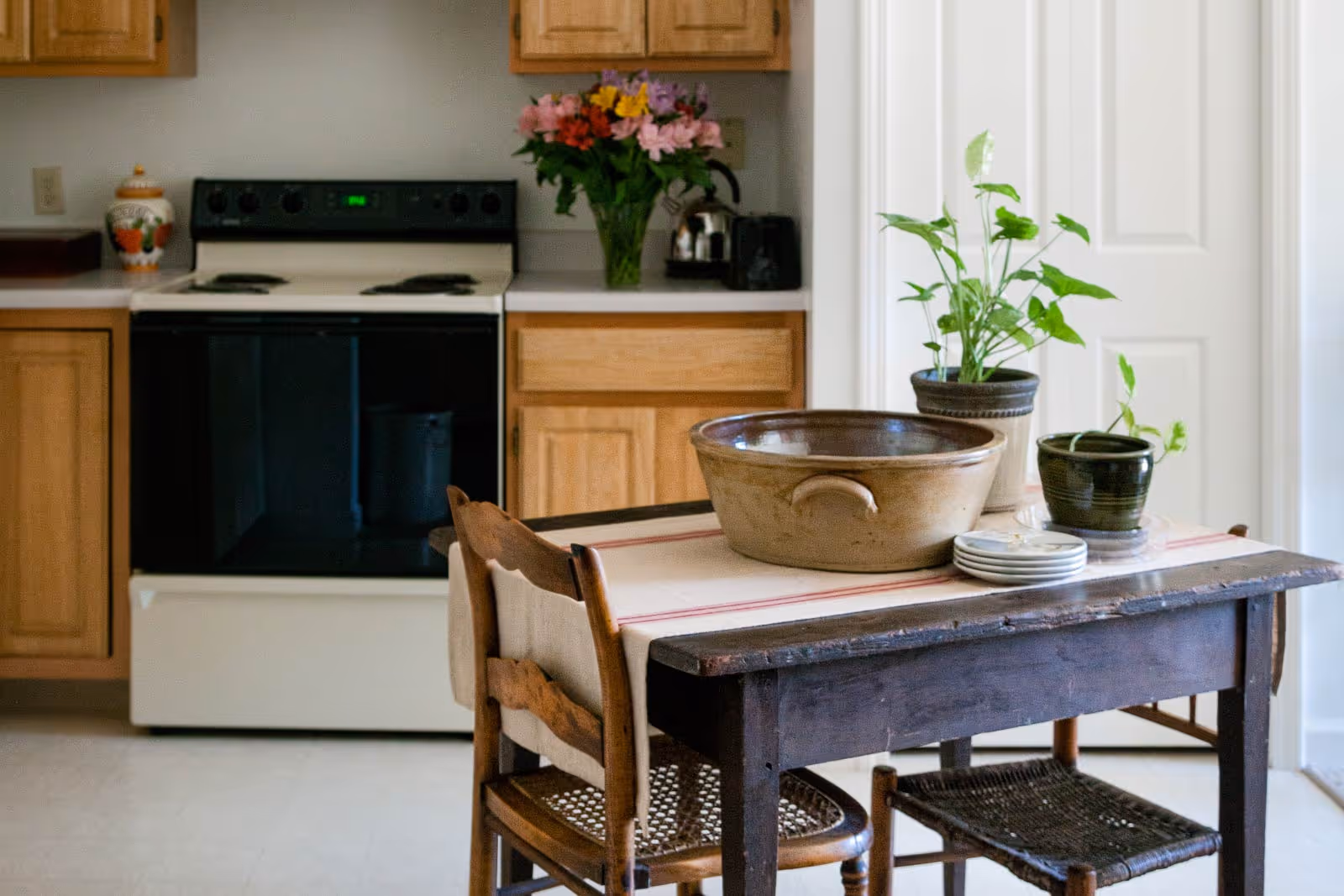 A cozy kitchen with wooden cabinets and a stove, and a small wooden table topped with bowls, plates and potted plants.