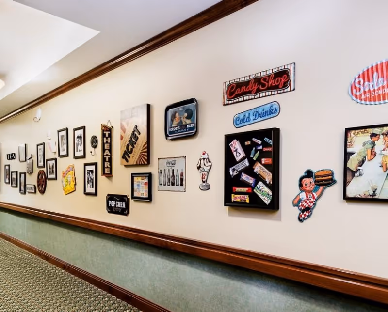 A hallway wall decorated with vintage-style signs and framed pictures, including themes of candy shop, theatre, popcorn, soda, and classic advertisements, with a patterned carpet and wooden trim along the wall.
