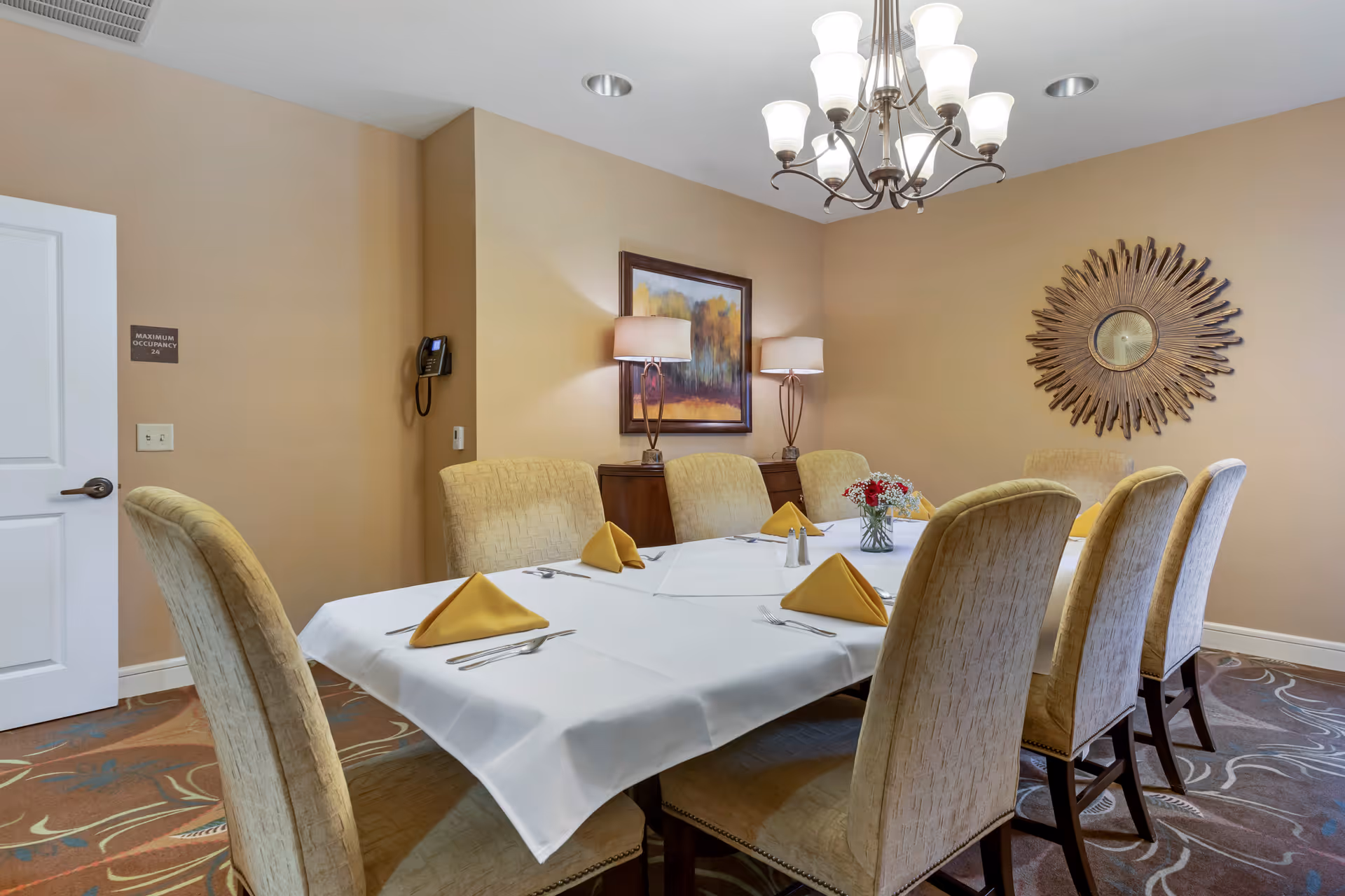A dining room with a rectangular table covered with a white tablecloth, set with yellow folded napkins, silverware, and a small flower arrangement in the center. The room has beige walls, a decorative sunburst mirror, a framed landscape painting, two table lamps, and eight upholstered chairs around the table.