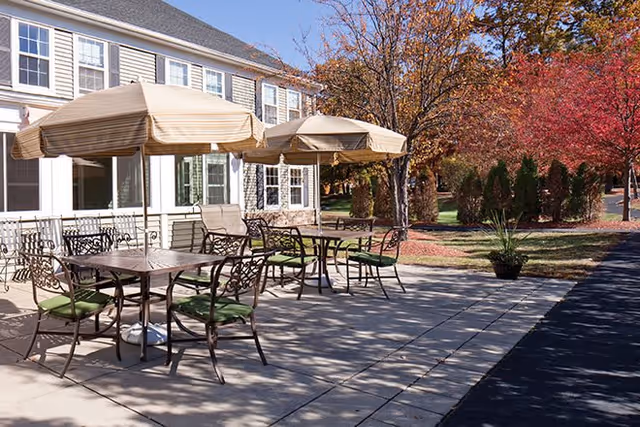 Outdoor patio area with metal tables and chairs featuring green cushions, shaded by beige umbrellas. The patio is adjacent to a building with multiple windows, surrounded by trees with autumn foliage and a paved walkway.